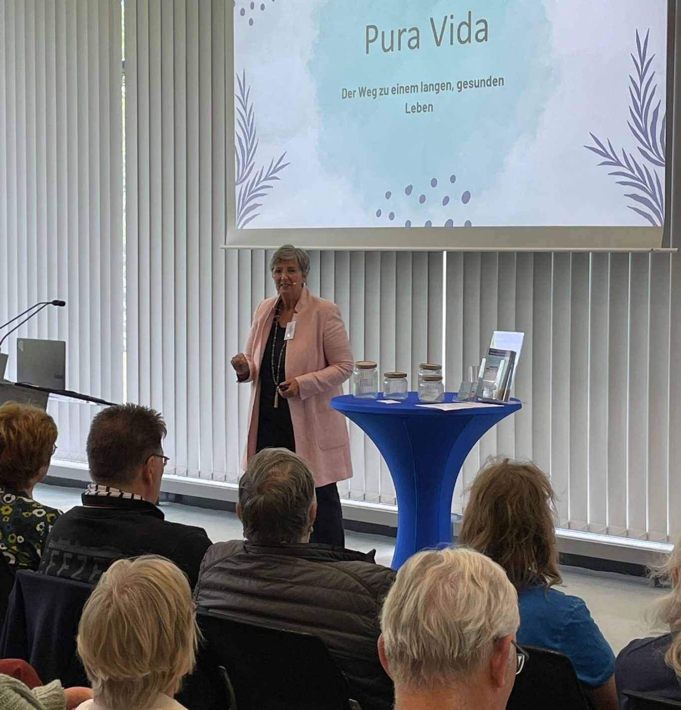 Volksbank-Mitgliedertag-2025-129.jpg Frau spricht an einem Tisch vor Publikum, im Hintergrund steht "Pura Vida".