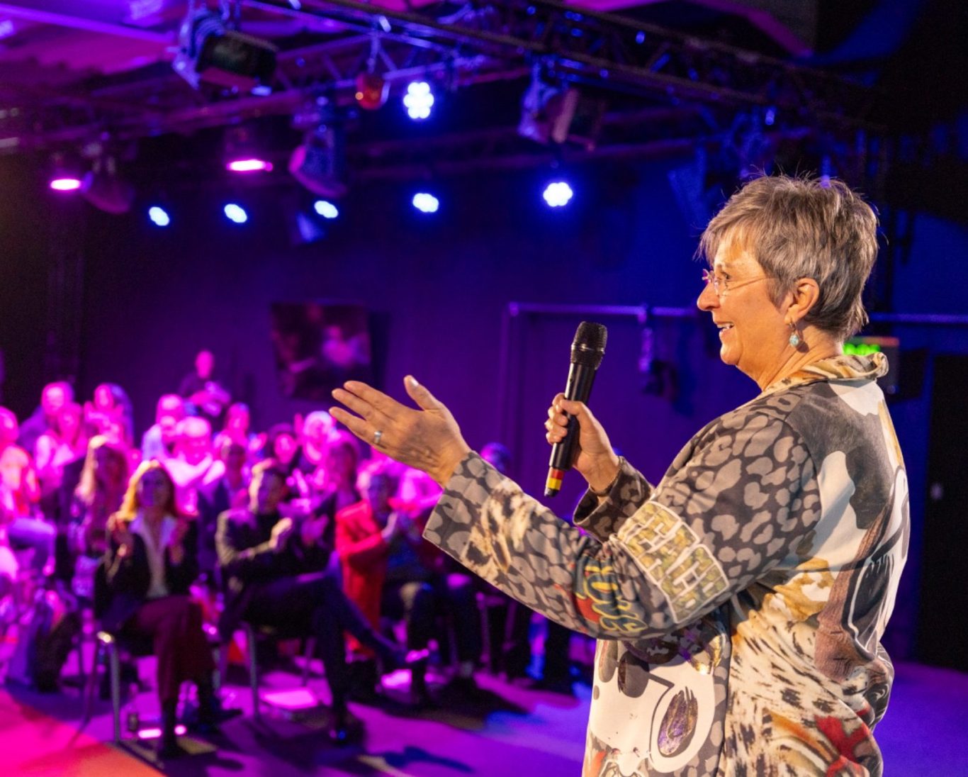 Monika Bröhl im Vortrag Eine Frau spricht mit einem Mikrofon vor einem Publikum in einem beleuchteten Raum.
